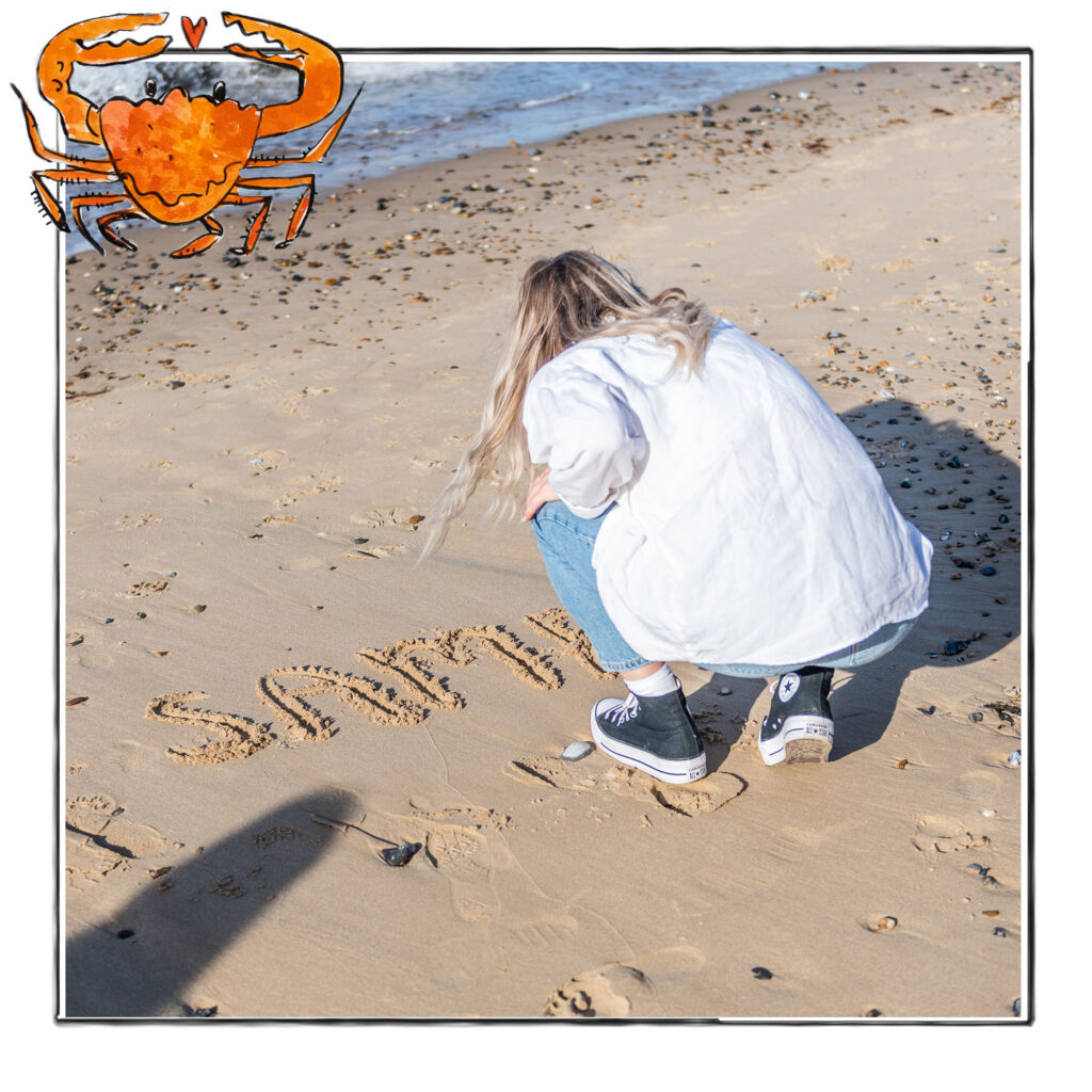 Image of a woman writing the word Samphire in the sand on the beach in Norfolk, UK, framed with a sketch line and overlaid with a hand drawn image of a crab